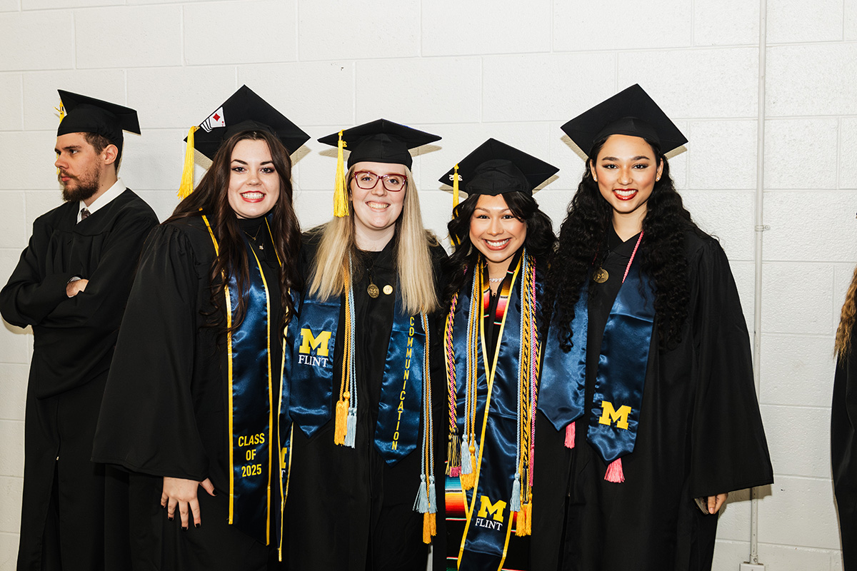 Five graduates in black caps and gowns pose together, wearing stoles that include "M FLINT" and "CLASS OF 2025."