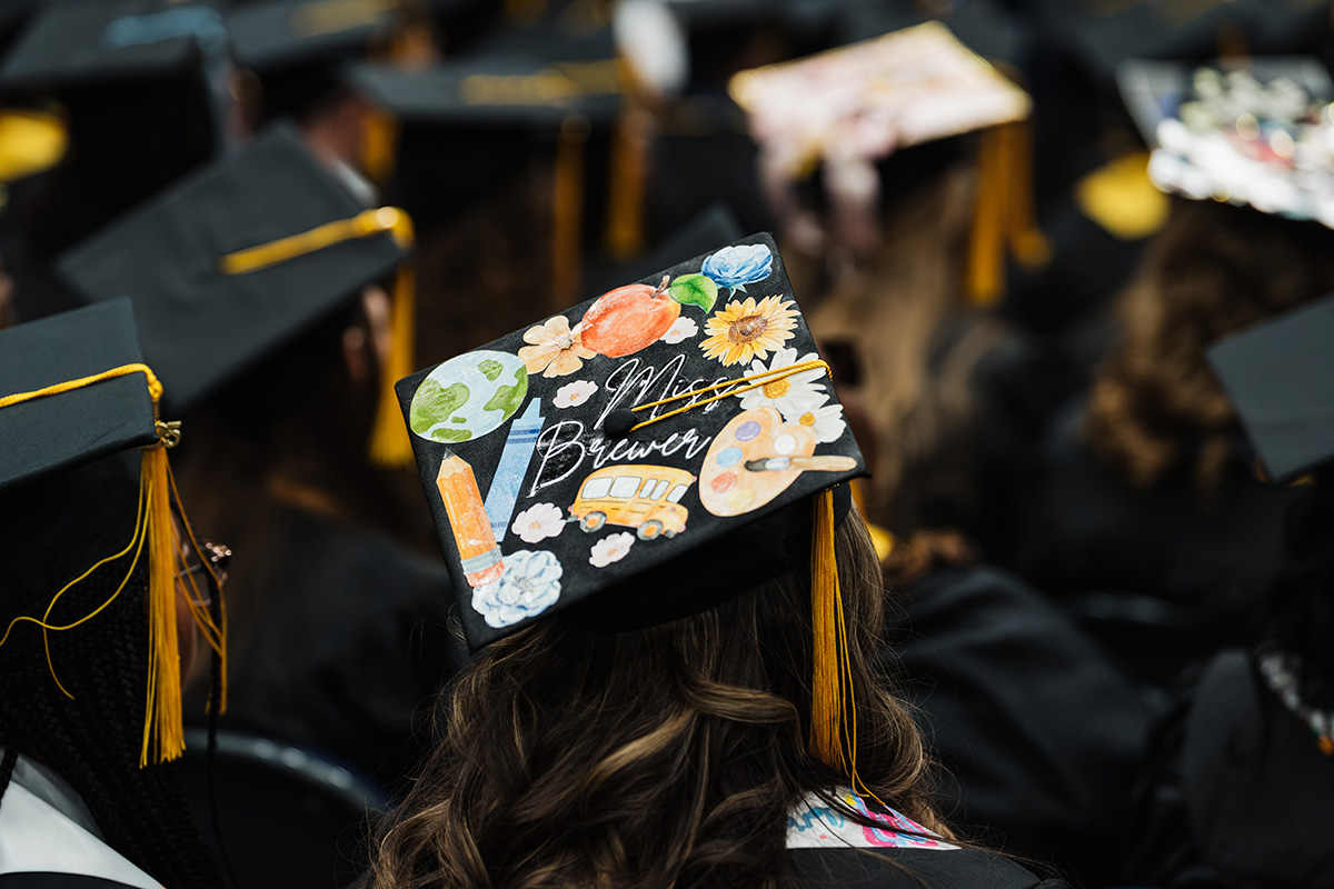 A decorated graduation cap featuring the name "Miss Brewer" along with colorful illustrations of a globe, flowers, a pencil, a bus, and a paint palette.