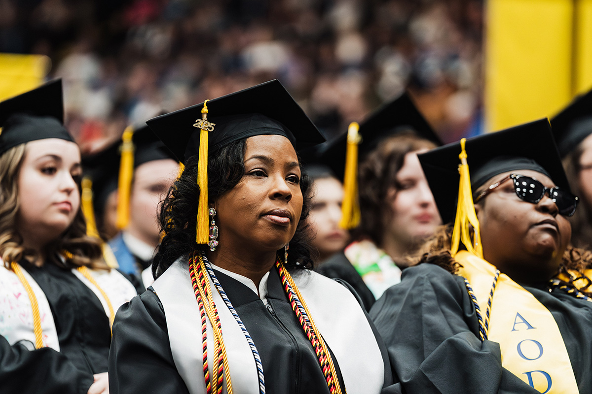 A group of graduates wearing black caps and gowns, with some adorned with colorful cords and stoles, seated in a ceremony.