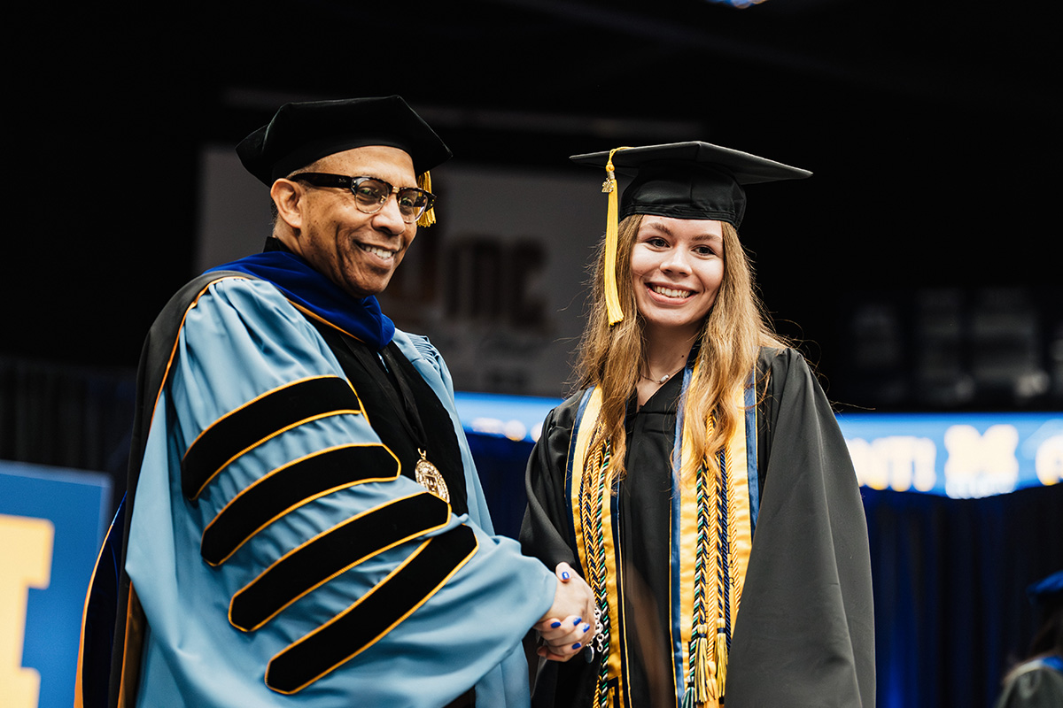 A graduate in a black cap and gown shakes hands with a person in academic regalia, set against a graduation ceremony backdrop.