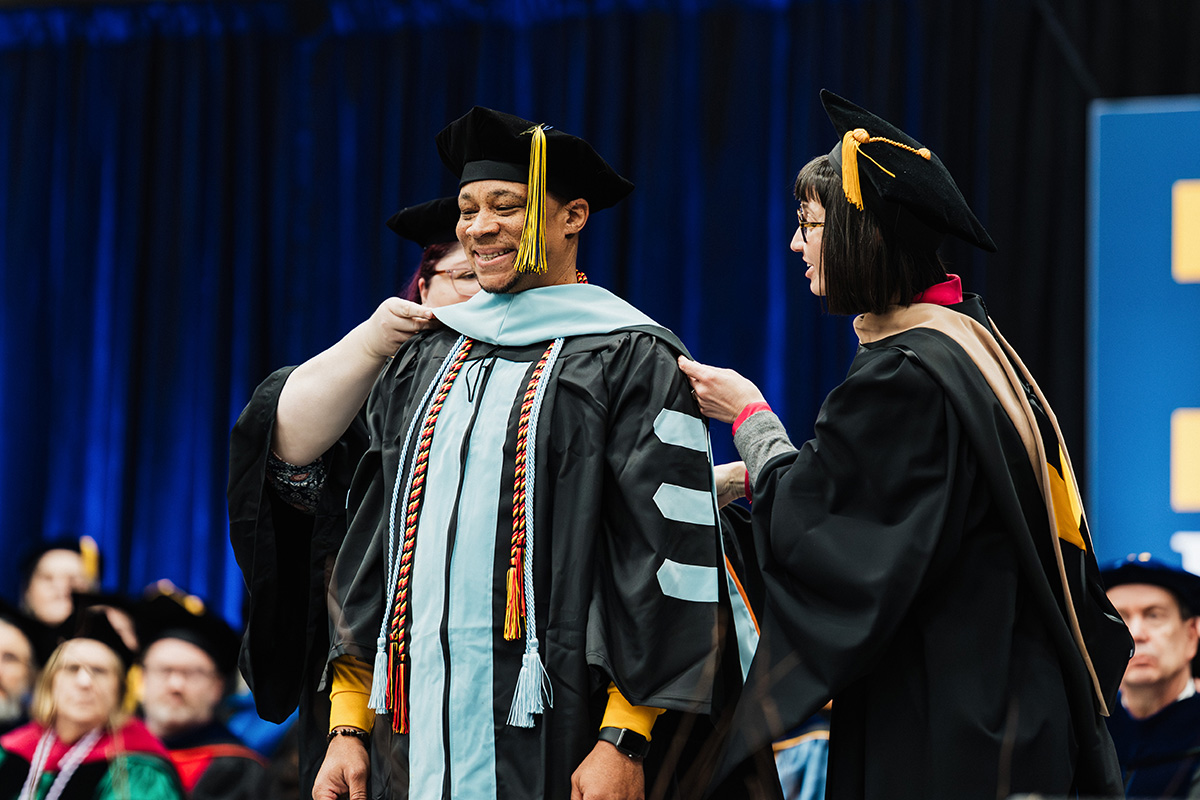 A person in academic regalia is receiving a hood from another individual during a graduation ceremony, with a blurred audience in the background.