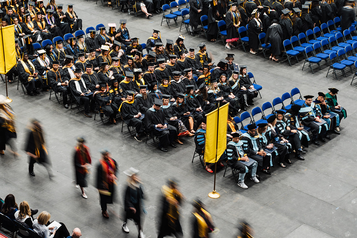 A graduation ceremony scene with rows of graduates in caps and gowns seated in blue chairs, some holding phones. A yellow banner is visible in the foreground, and individuals in academic regalia are walking in the background.