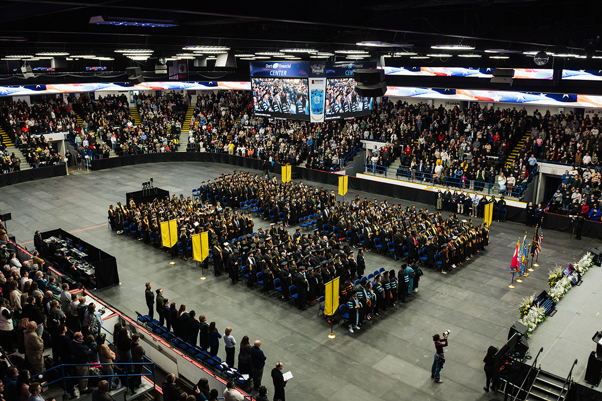 A large graduation ceremony taking place in an arena, with graduates in caps and gowns seated in rows. The audience is standing and applauding, and there are flags and banners displayed. A large screen shows the event, and flowers are arranged on the stage.