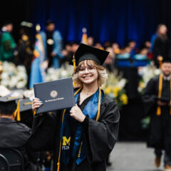 A graduate in a black cap and gown holds a diploma from the University of Michigan-Flint, smiling at the camera during a graduation ceremony. In the background, other graduates and faculty members are visible.