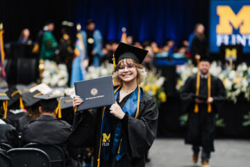 A graduate in a black cap and gown holds a diploma from the University of Michigan-Flint, smiling at the camera during a graduation ceremony. In the background, other graduates and faculty members are visible.