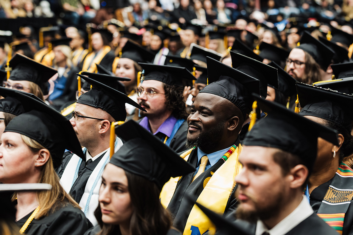 A crowd of graduates wearing black caps and gowns, some with colorful stoles, seated in a large auditorium during a graduation ceremony.
