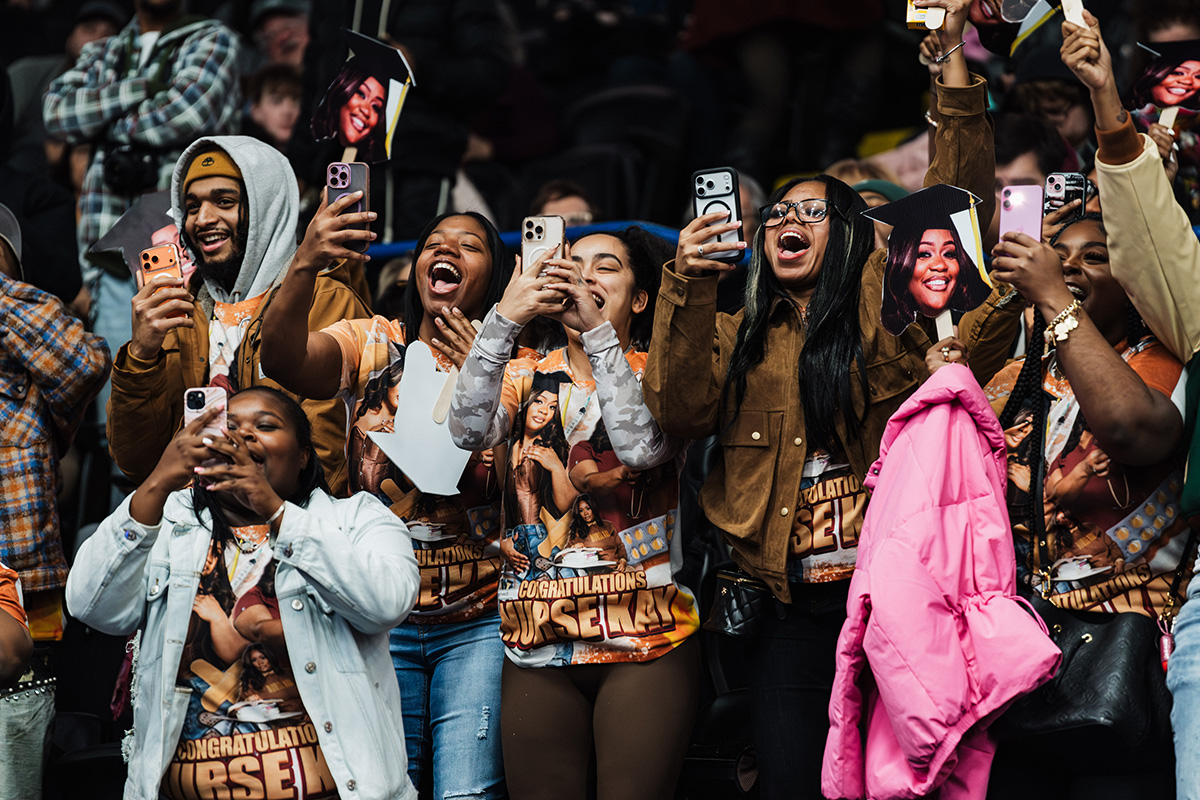A group of people wearing matching shirts with a celebratory design, holding up smartphones to capture a moment at an event.