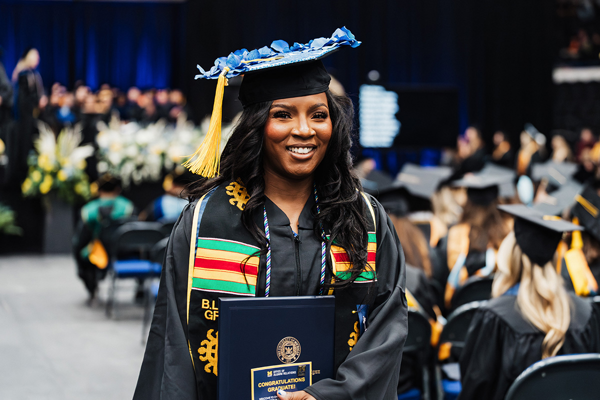 A graduate wearing a cap and gown holds a diploma, walking across a stage decorated with flowers, while other graduates are seated in the background.