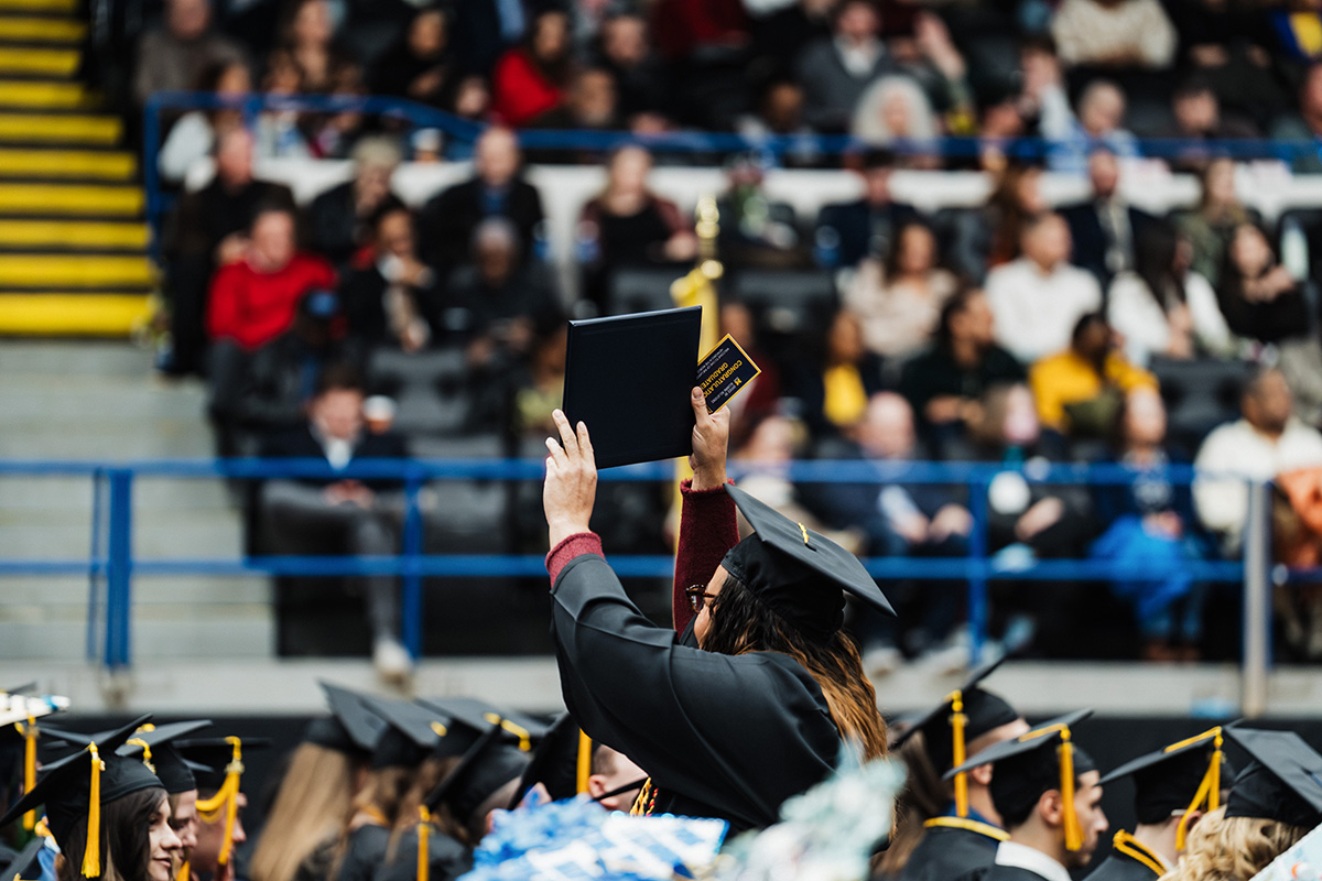 A graduate in a black cap and gown holds up a diploma during a graduation ceremony, with an audience in the background.
