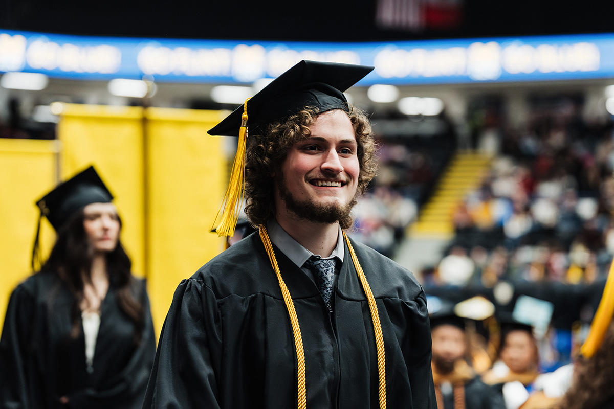 A graduate wearing a black cap and gown with a gold tassel stands in a crowded auditorium, with yellow banners in the background.