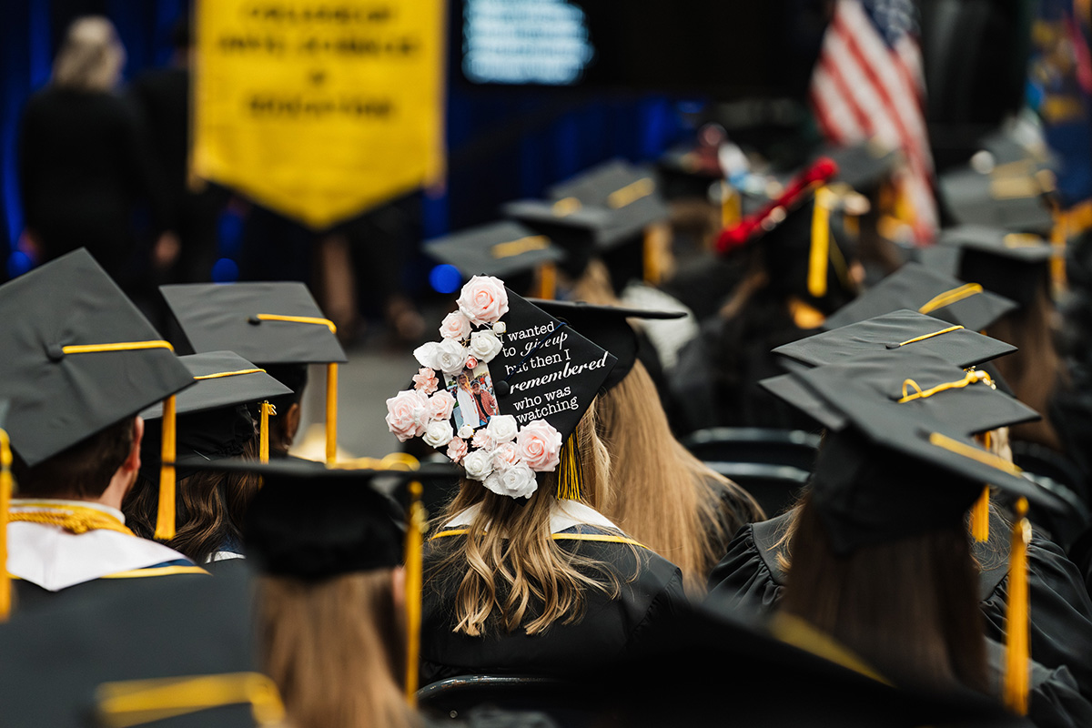 A close-up of a graduation cap decorated with white flowers and the text: "I wanted to give up, but then I remembered who was watching." The cap is worn by a graduate sitting among others in black caps and gowns.
