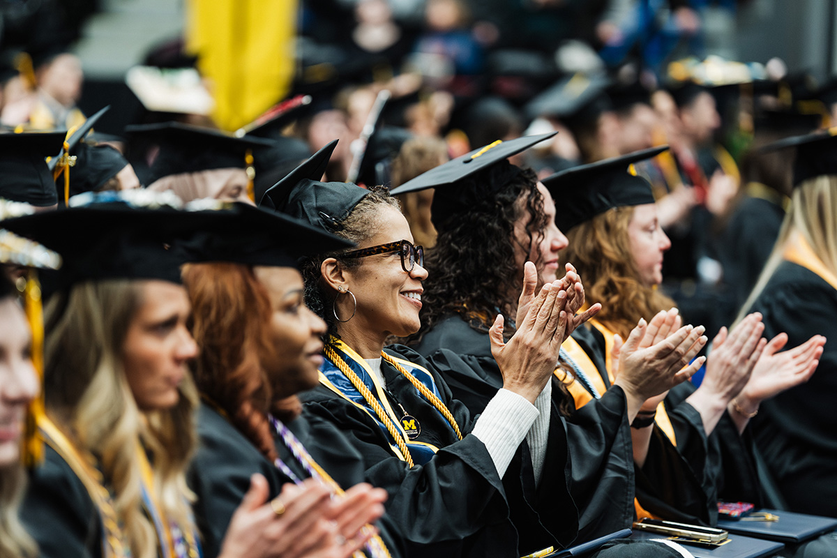 A group of graduates in black caps and gowns, some wearing honor cords, are seated and applauding during a ceremony.