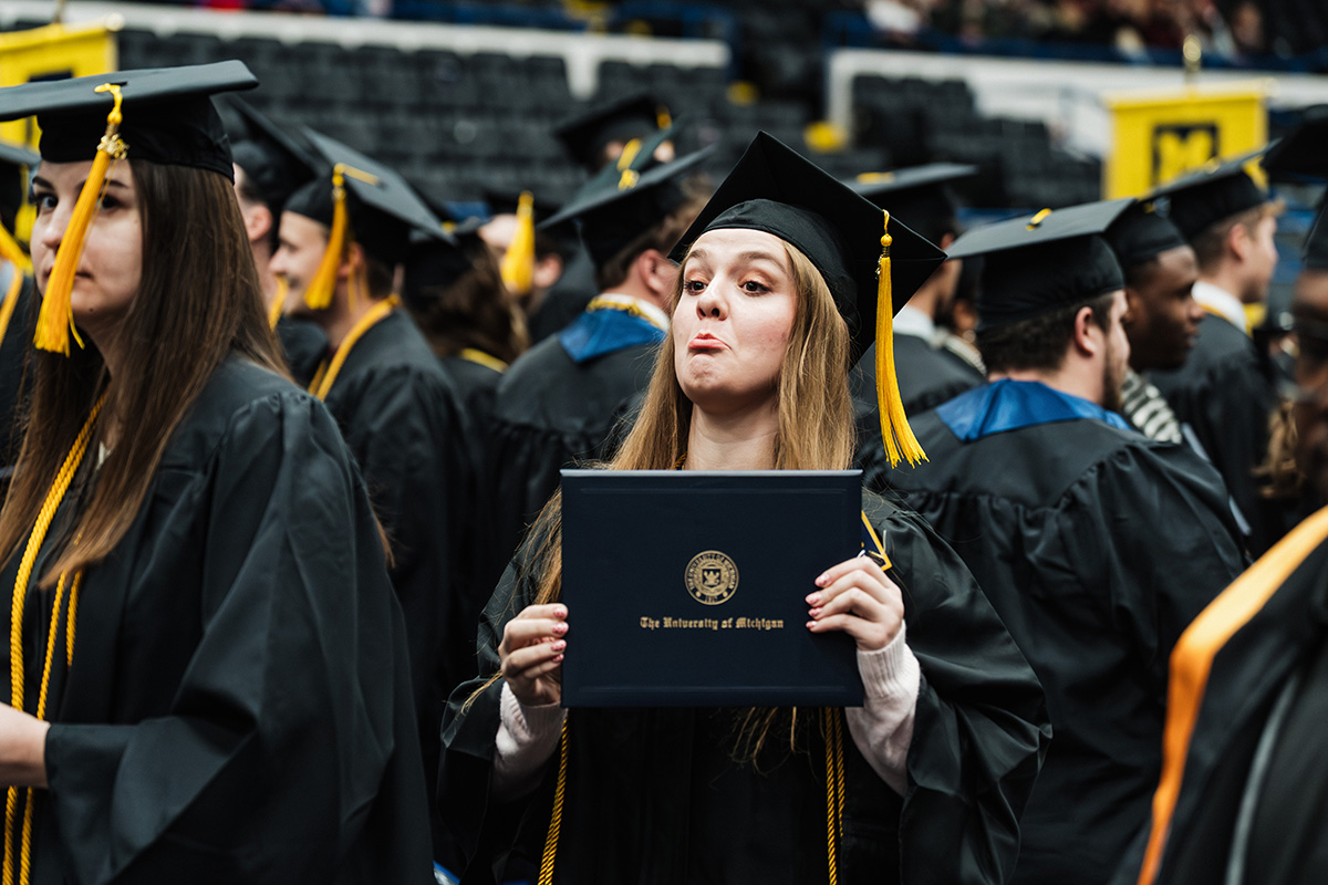 A graduate in a black cap and gown holds a diploma from the University of Michigan during a graduation ceremony, surrounded by fellow graduates.