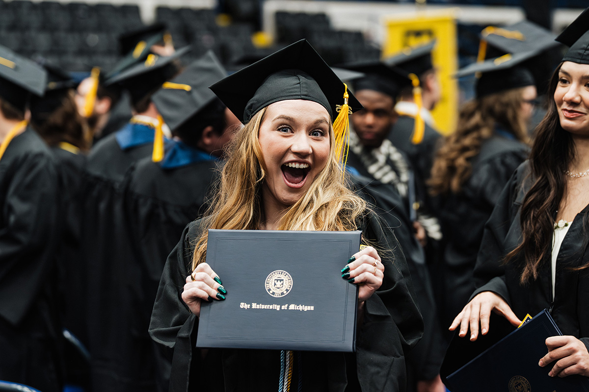 A graduate in a black cap and gown holds a diploma that reads "The University of Michigan" while surrounded by fellow graduates.