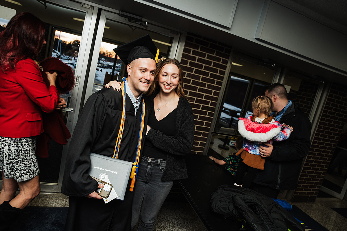 A graduate in a black cap and gown holds a diploma and poses for a photo with a woman. In the background, a man holds a child, and another person is partially visible. The setting appears to be a school or event venue.