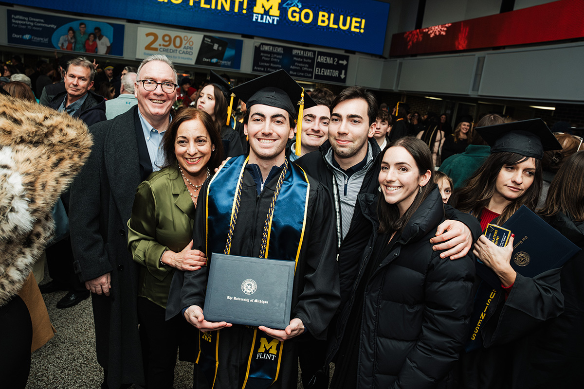 A group of people celebrating a graduation, with one person in a cap and gown holding a diploma from the University of Michigan Flint. A large banner in the background reads "GO FLINT! GO BLUE!"