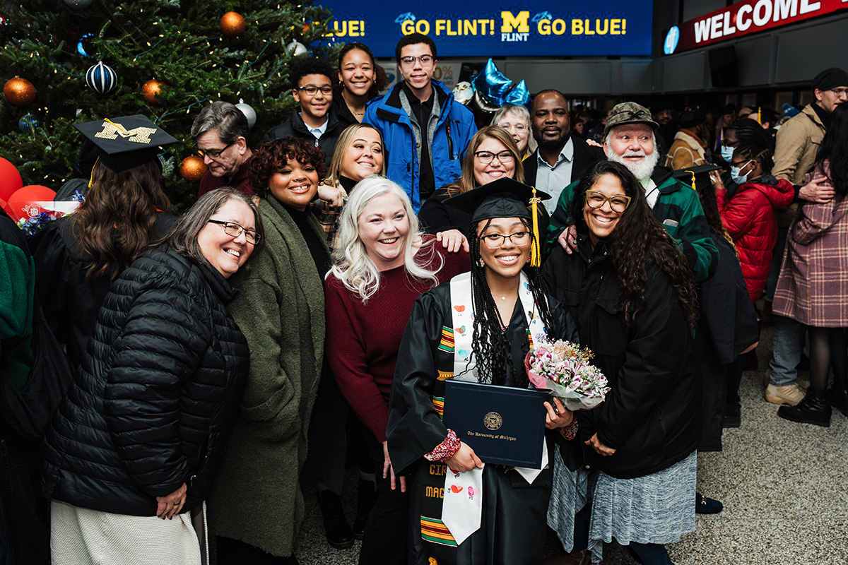 A group of people gathered together, celebrating a graduation. One person in a cap and gown holds a diploma and flowers. In the background, a decorated Christmas tree and a sign that reads "GO FLINT! GO BLUE!" are visible.