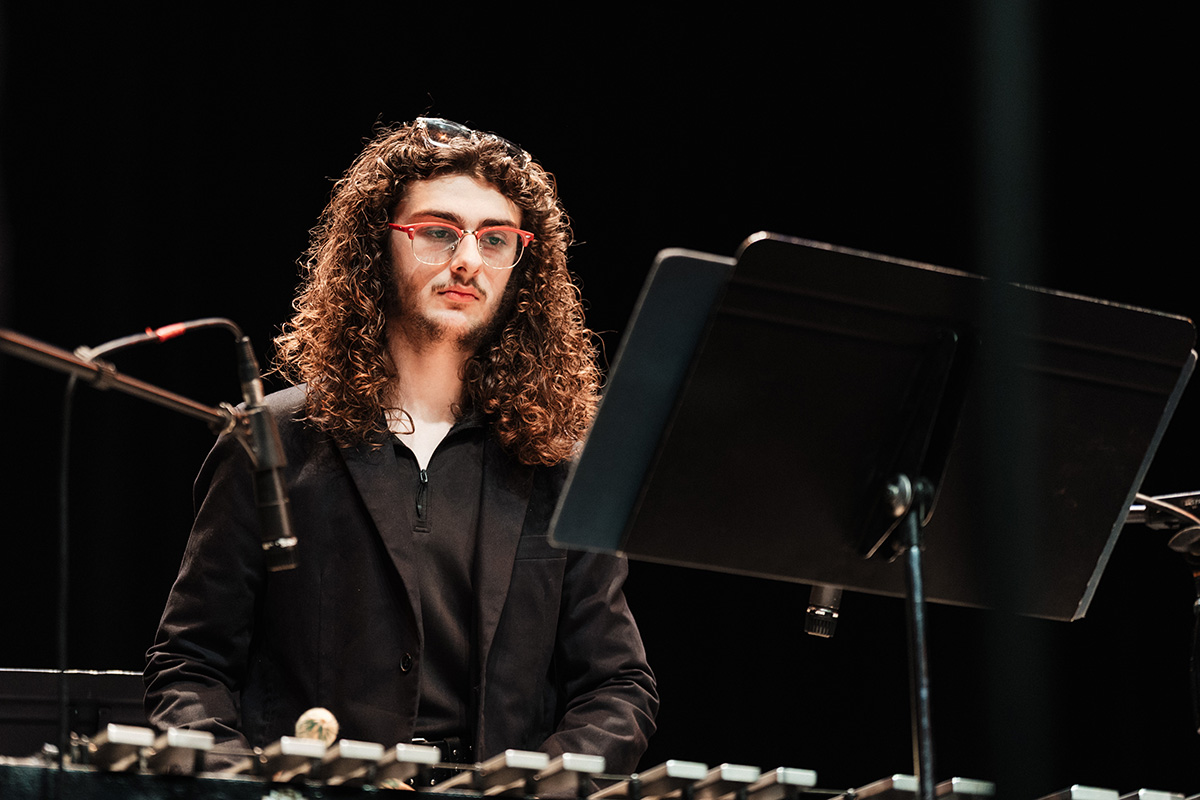 A musician with curly hair plays a vibraphone, with music stands in the background.