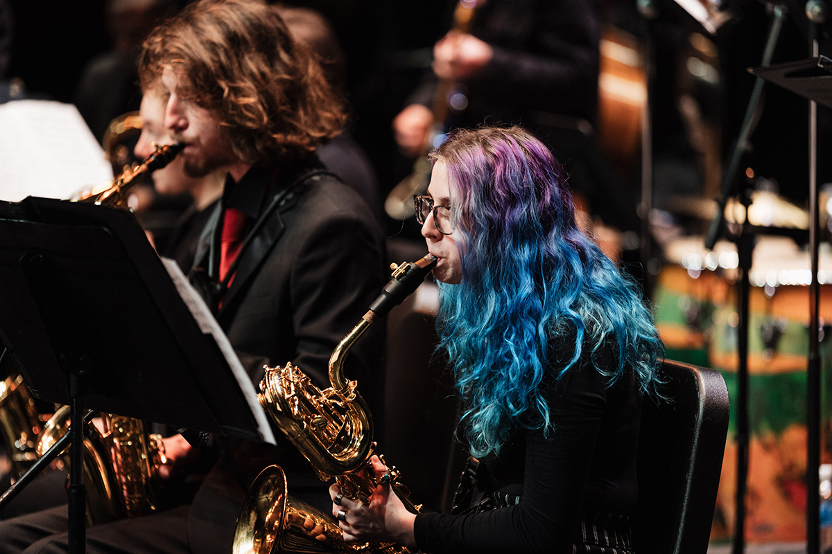 A musician with blue and purple hair plays a saxophone in an orchestra setting, with other musicians in the background.