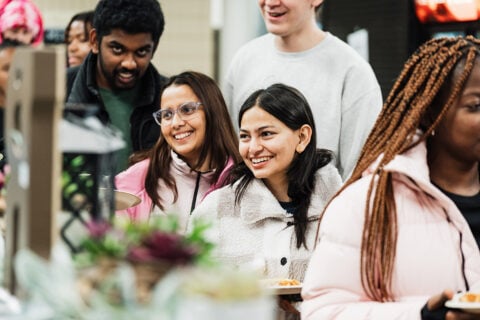 A group of people standing in line, some holding plates of food, in a casual dining setting.