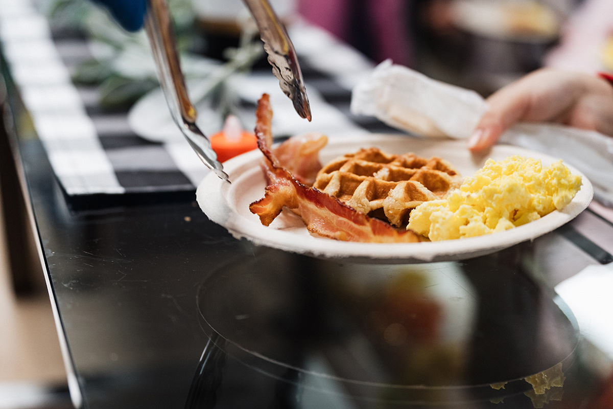 A plate with bacon, waffles, and scrambled eggs being served.