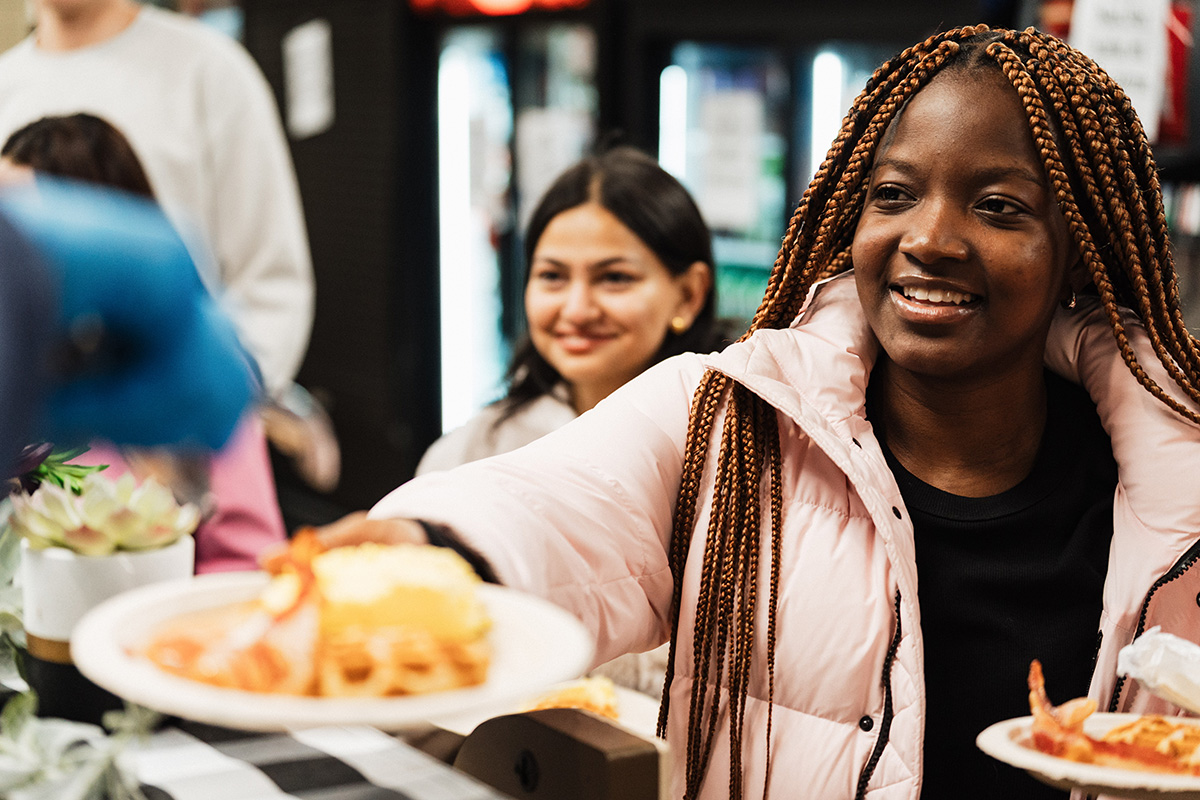 A person wearing a pink jacket is holding a plate of food, including pasta and shrimp, in a dining setting.