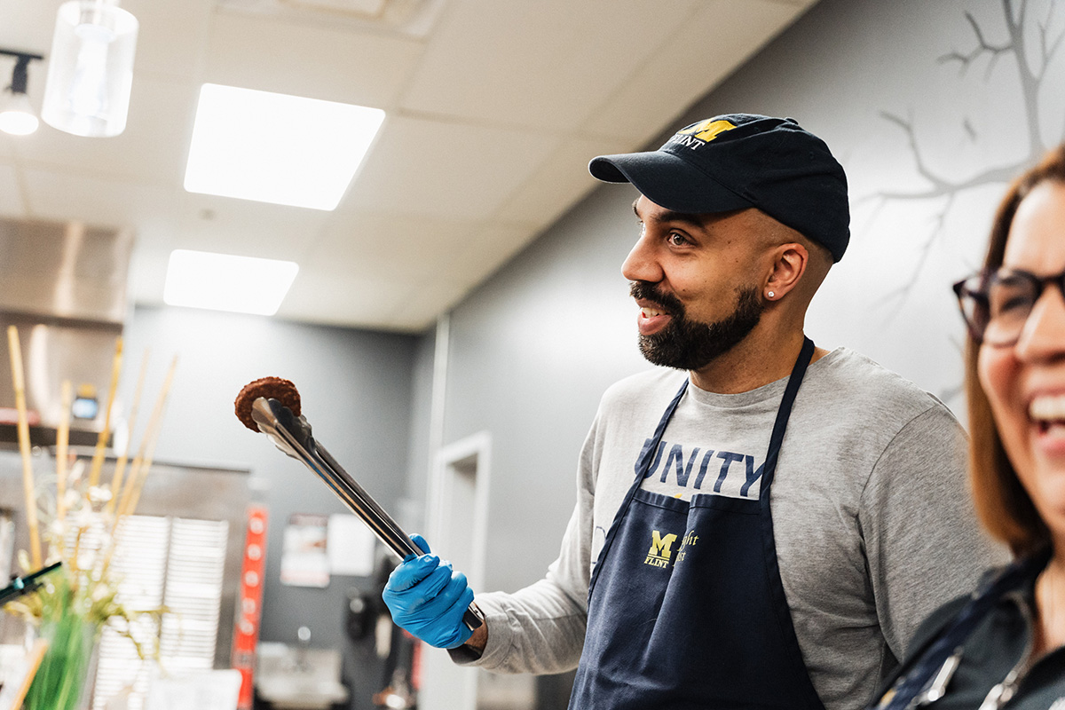 A person wearing a blue apron and gloves holds tongs, smiling while interacting with another person in a kitchen setting.
