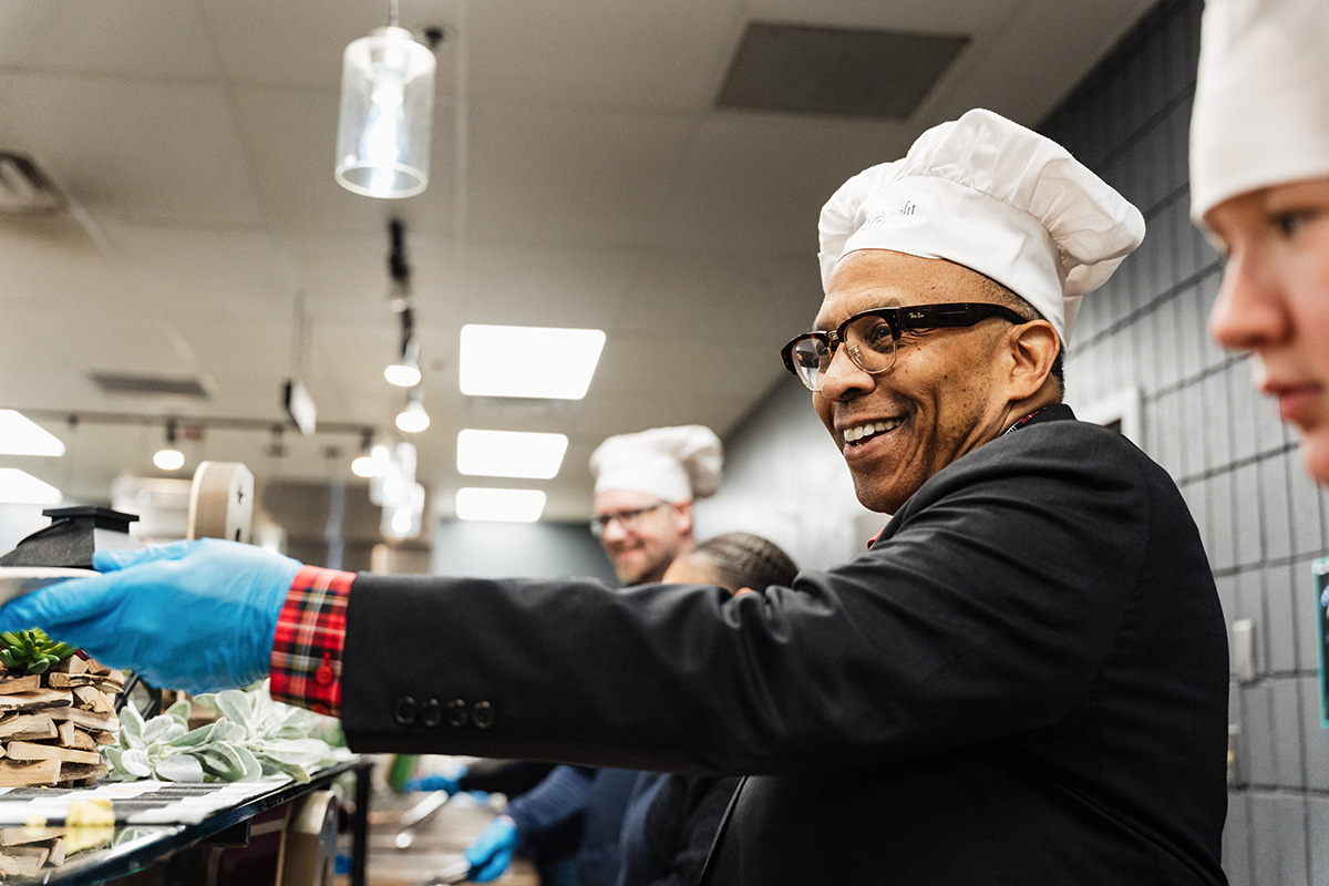 A chef in a black jacket and white chef's hat gestures while preparing food, wearing blue gloves in a kitchen setting.