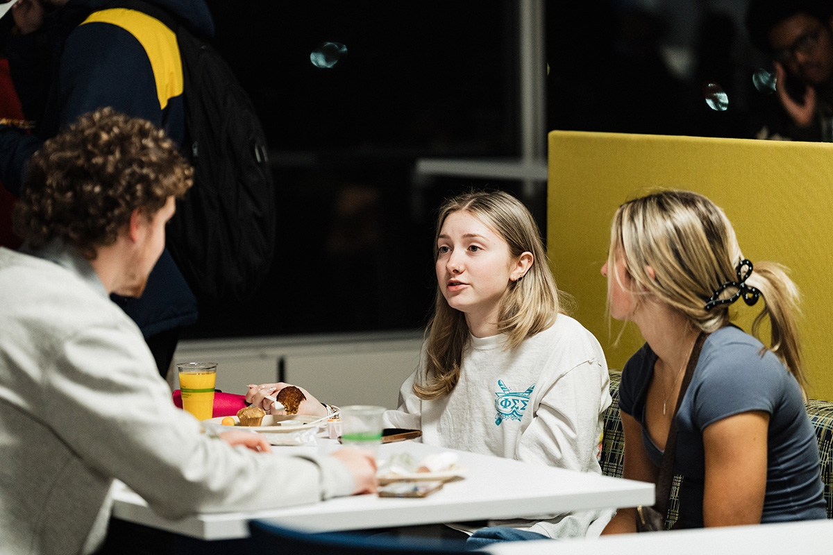 Three people are seated at a table with food and drinks, engaged in conversation in a dimly lit setting.
