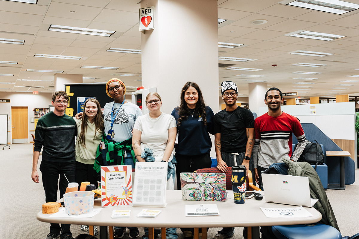 A group of six people standing in front of a table with various items, including a sign that says "Save the Superheroes!" and a laptop.