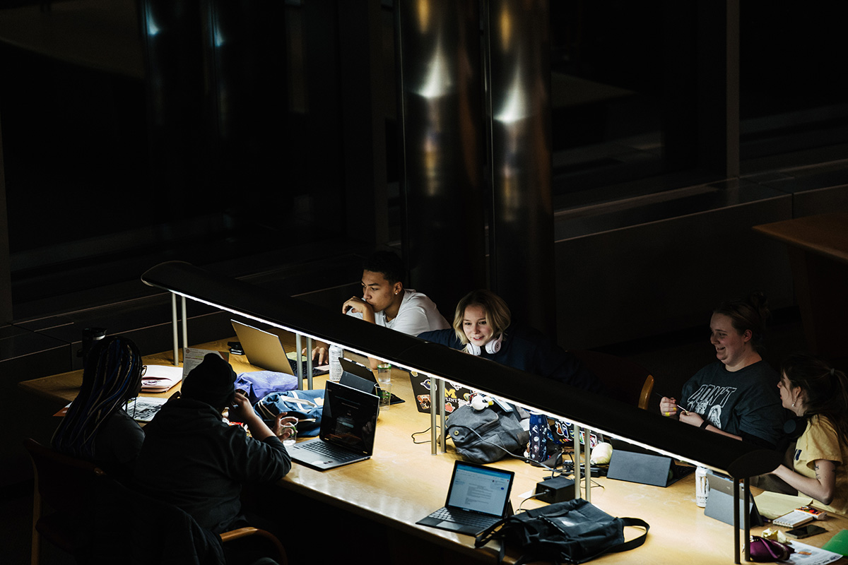A group of people working at a table with laptops and personal items in a dimly lit environment.