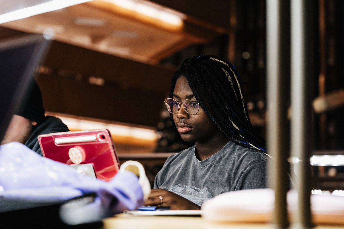 A person with braided hair is sitting at a table, using a tablet with a red case, in a study environment.