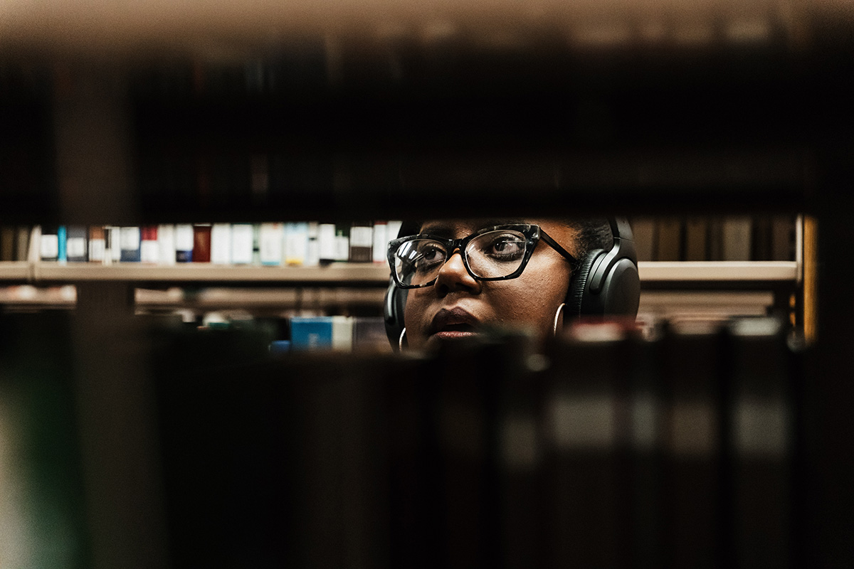 A person wearing headphones is seated at a table in a library, with bookshelves in the background.