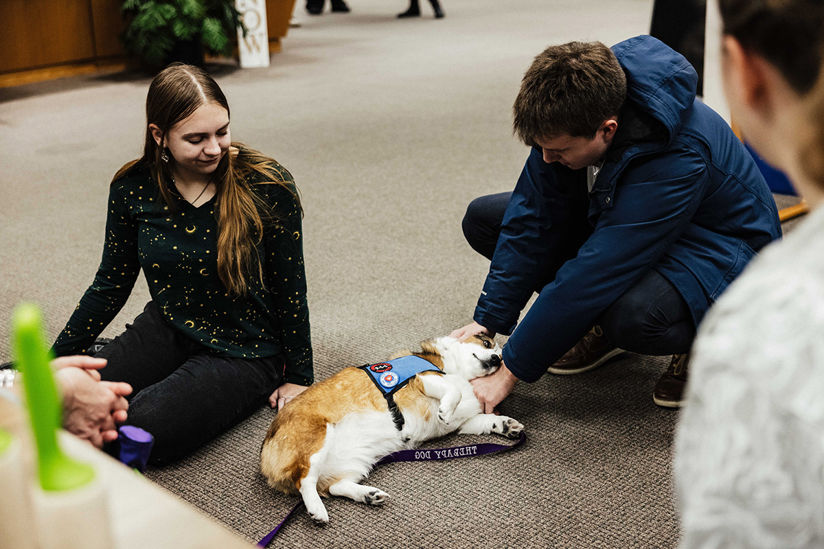 A person in a blue jacket gently interacts with a relaxed dog wearing a service vest, while another person sits nearby.