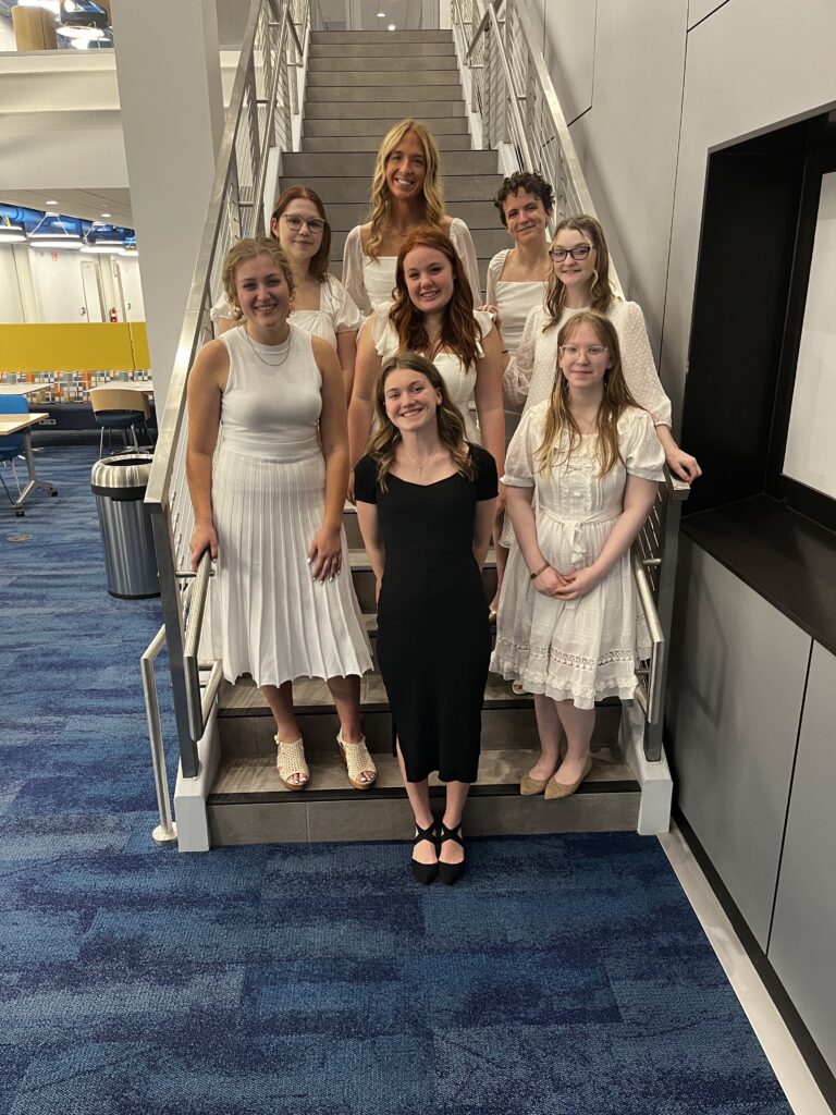 A group of seven women in white and black dresses poses on a staircase in a modern indoor setting.