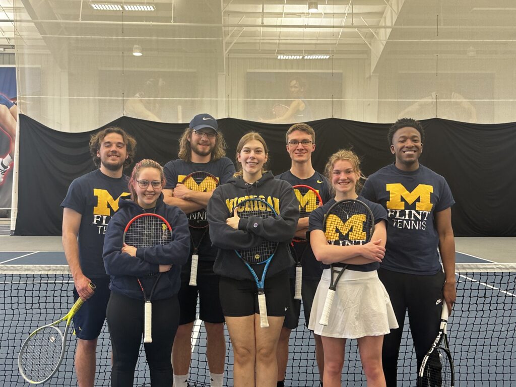 A group of seven tennis players posing together on a court, wearing matching shirts with "Flint Tennis" and "Michigan" logos.