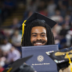A graduate wearing a cap and gown holds a diploma from the University of Michigan during a graduation ceremony.