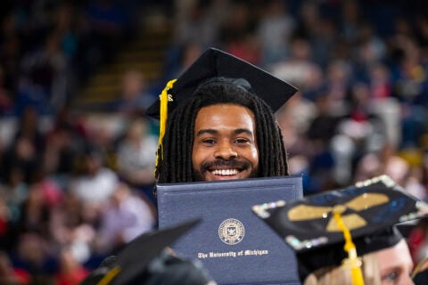 A graduate wearing a cap and gown holds a diploma from the University of Michigan during a graduation ceremony.