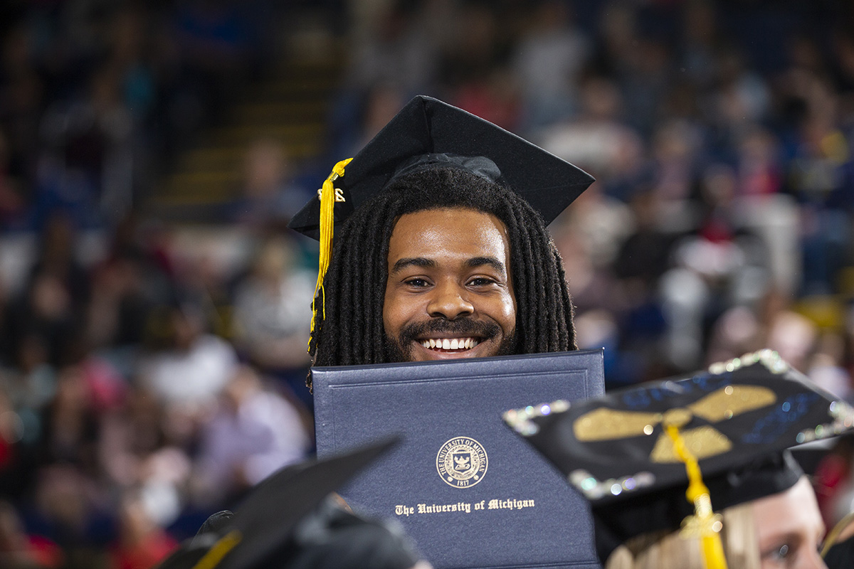 A graduate wearing a cap and gown holds a diploma from the University of Michigan during a graduation ceremony.