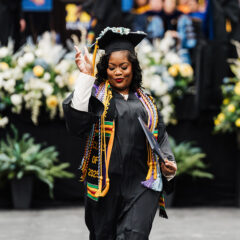A graduate in a cap and gown holds a diploma and makes a peace sign, with flowers and graduates in the background.