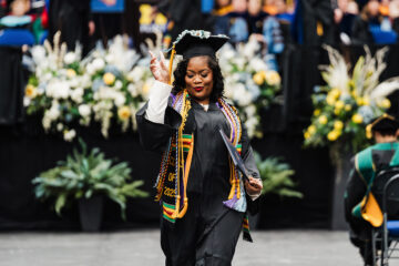 A graduate in a cap and gown holds a diploma and makes a peace sign, with flowers and graduates in the background.