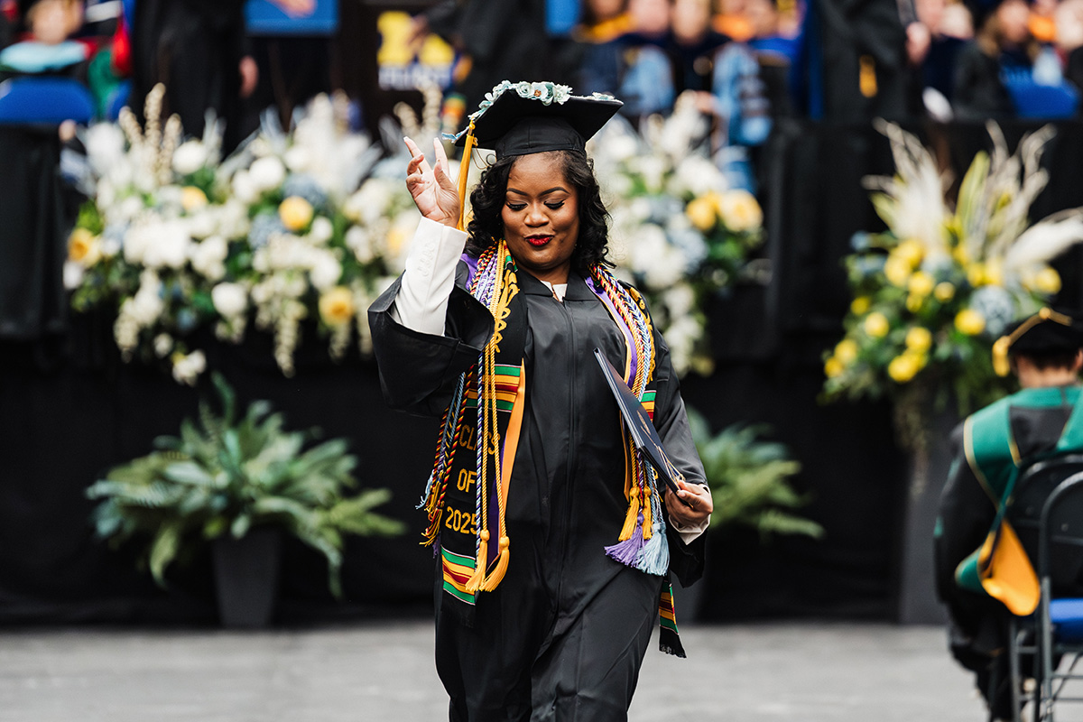 A graduate in a cap and gown holds a diploma and makes a peace sign, with flowers and graduates in the background.