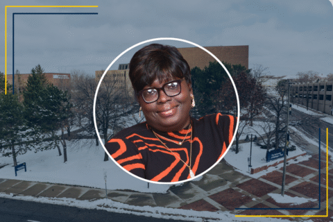 A circular portrait of a person wearing a black and orange striped sweater, with a snowy campus background.