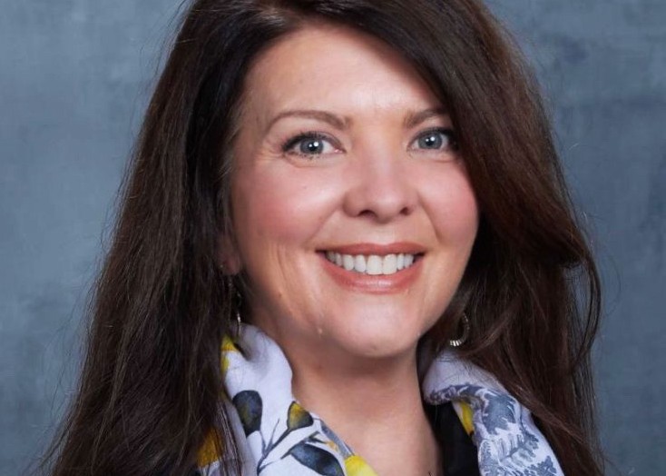 A woman with long brown hair wearing a patterned scarf, set against a gray background.