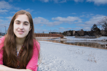 A person with long hair wearing a pink shirt is sitting on a snowy bank, with a wooden bridge and a river in the background under a blue sky with clouds.