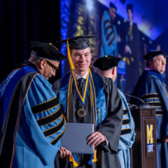 A graduate in a cap and gown receives a diploma from a faculty member during a graduation ceremony, with other graduates and faculty in the background.