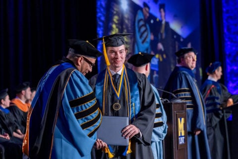 A graduate in a cap and gown receives a diploma from a faculty member during a graduation ceremony, with other graduates and faculty in the background.
