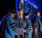 A graduate in a cap and gown receives a diploma from a faculty member during a graduation ceremony, with other graduates and faculty in the background.