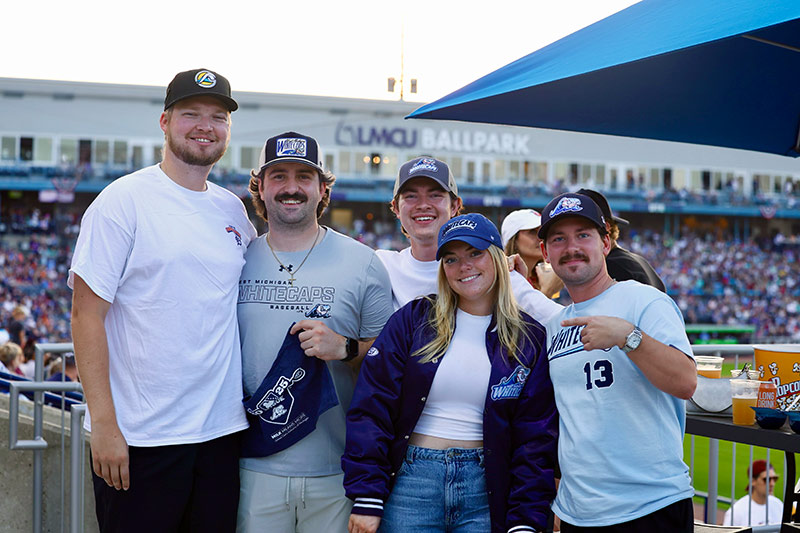 A group of five people posing together at a baseball game, wearing team hats and apparel, with a stadium in the background.
