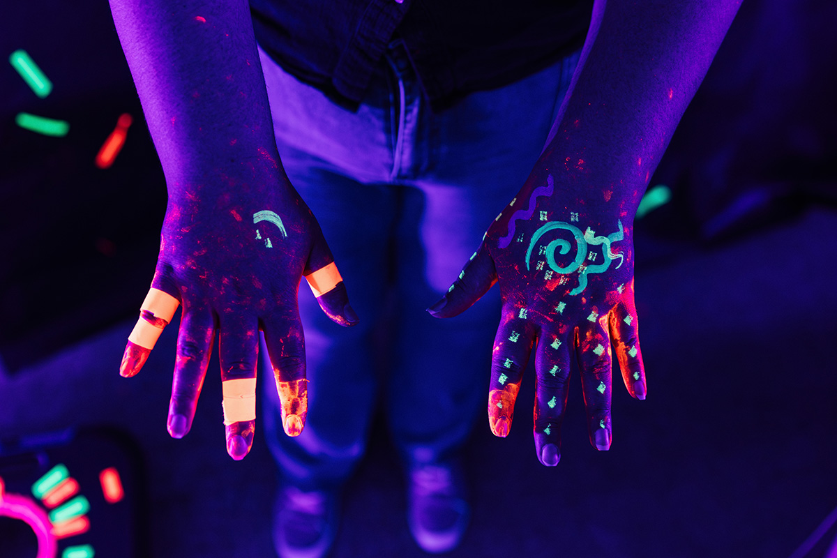 FLINT_Light Up the Night 2026_0003 Hands covered in colorful glow-in-the-dark paint, with various patterns and bright tape on the fingers, against a dark background.